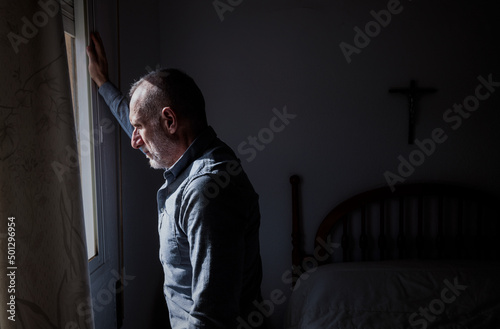 Adult man looking through window in bedroom with religious cross on wall © WeeKwong