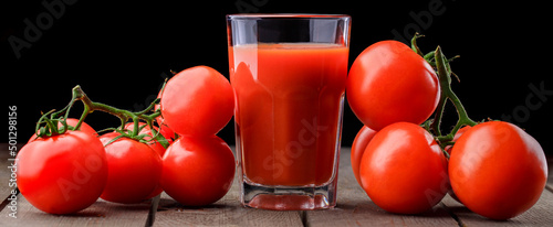 A glass cup with tomato juice, next to it lies a branch of tomatoes. On a wooden table there is a glass of tomato juice and tomatoes lie on a black background.