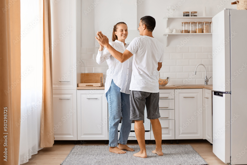 Indoor shot of wife and husband wearing casual clothing dancing together in kitchen, holding hands, smiling, expressing positive emotions and romantic feelings. Indoor shot of wife and husband wearing casual clothing dancing together in kitchen, holding hands, smiling, expressing positive emotions and romantic feelings.