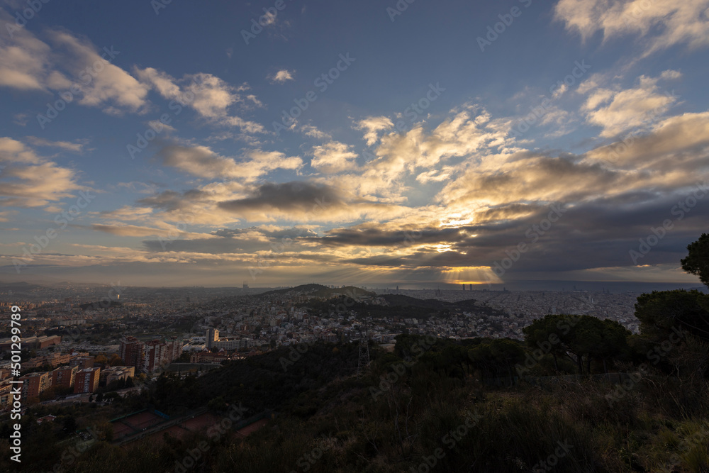 Picturesque landscape of Barcelona from the hill in the early morning. Sunbeams through the clouds. Dramatic sky over the city. Autumn in Barcelona, Spain.
