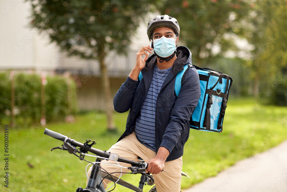 food shipping, health and people concept - indian delivery man in bike helmet and mask with thermal insulated bag and smatphone riding bicycle on city street