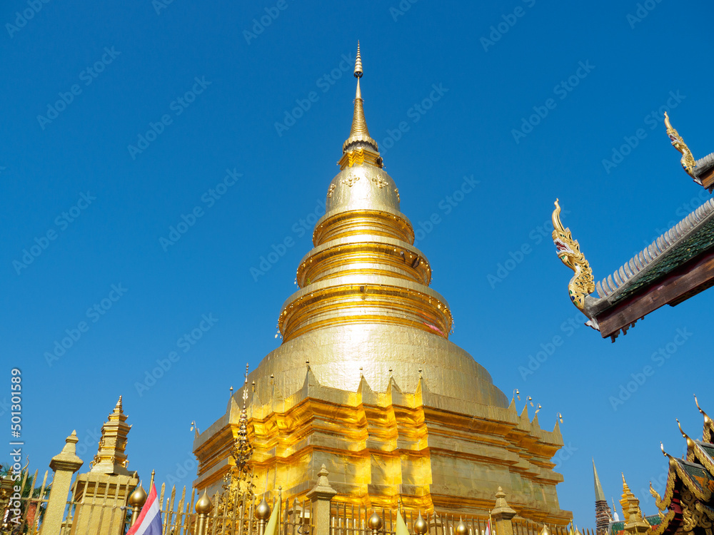 Naklejka premium Phra Maha That Chedi, ancient pagoda at Wat Phra That Haripunchai, Lamphun, Thailand. The pagoda beautifully covered with Django gold, against bright blue sky background.