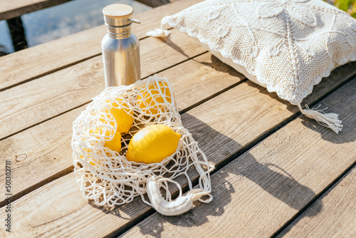 Yellow lemons and water metal bottle in the eco friendly net bag at wooden pearce at summer outdoor. Eco modern shopper with citrus. Zero waste concept.