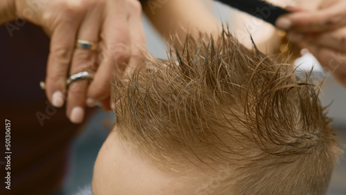 Blond caucasian boy is sitting in barber shop and hairdresser girl is cutting his hair. Child gets fashionable haircut, hairdresser cuts child hair with scissors. Close up of hairdresser cutting hair.