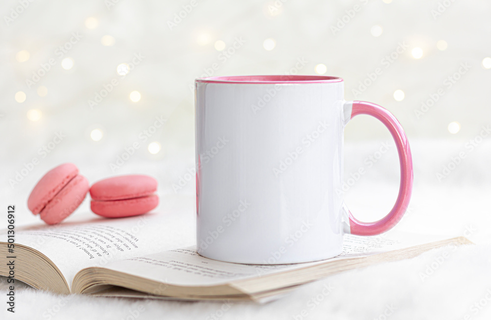 Blank pink handle mug on the book with white bokeh lights and pink macaroons. Blank pink handle mug on the book with white bokeh lights and pink macaroons.