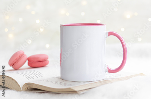 Blank pink handle mug on the book with white bokeh lights and pink macaroons.