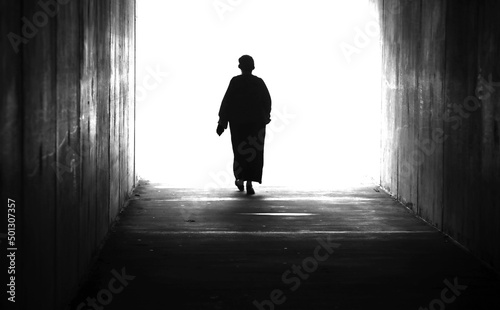 A black and white high contrast silhouette of figure in a landscape of a single lady or woman stepping or walking through a tunnel into and twowards a bright light.
