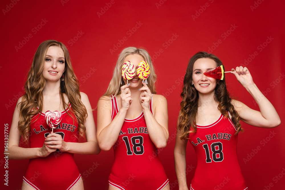 Beautiful and happy female friends in bright red swimsuits with bright candy lollipops in their hands on a red background, studio. Beautiful and happy female friends in bright red swimsuits with bright candy lollipops in their hands on a red background, studio.