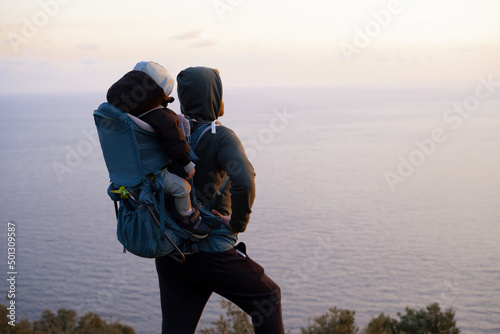 Young caucasian man hiking near beautiful blue sea carrying baby in bacpack,sling,carrier.Family travelling,trips,lifestyle.Father and child,infant,toddler spending time in nature.Copy space