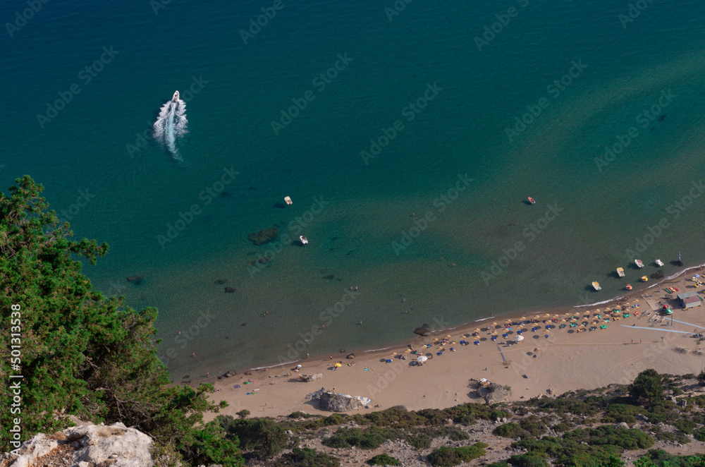 Fragment of the popular Tsampika beach with a boat in the azure sea and sun beds on the sandy shore top view (Rhodes, Greece) Fragment of the popular Tsampika beach with a boat in the azure sea and sun beds on the sandy shore top view (Rhodes, Greece)