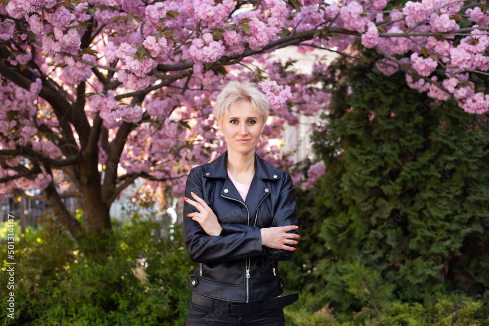 Young beautiful woman near sakura blooming tree. Pink flowers, spring and youth concept. Stylish girl smiling. Blooming garden with cherry-tree. Copyplace, place for text. Young beautiful woman near sakura blooming tree. Pink flowers, spring and youth concept. Stylish girl smiling. Blooming garden with cherry-tree. Copyplace, place for text.