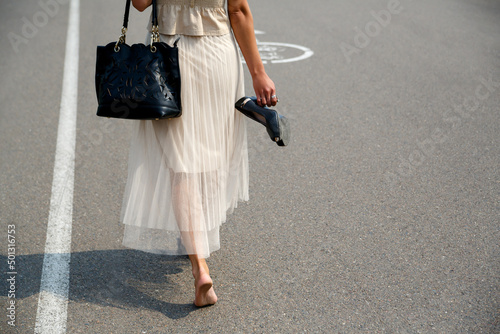 cropped image of bare dirty feet. The woman took off her shoes, holds the shoes in her hands, and walks barefoot along the asphalt road