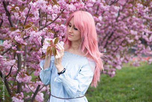 beautiful hipster woman in pink wig near sakura blooming tree. Pink flowers  spring and youth concept. Stylish girl smiling. Blooming garden with cherry-tree. Copyplace  place for text.