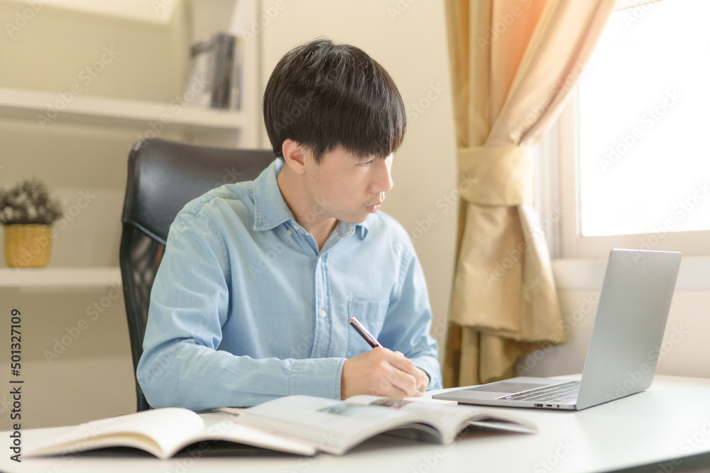 Young Asian college male student writing notes, using laptop for learning online education, business learning, watching online webinar. Focused man doing homework. Online education learning concept. Young Asian college male student writing notes, using laptop for learning online education, business learning, watching online webinar. Focused man doing homework. Online education learning concept.
