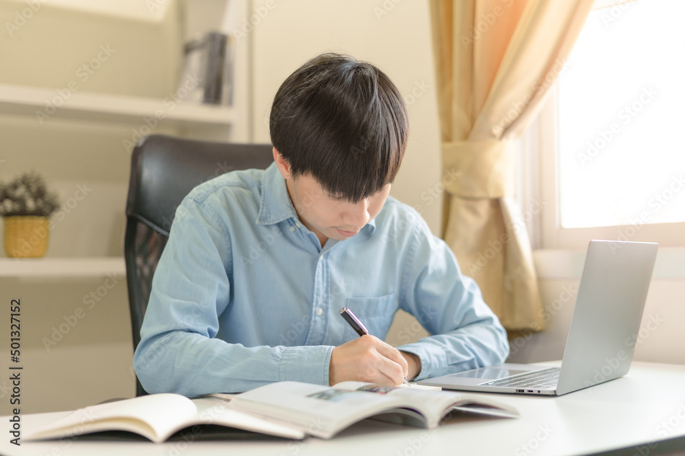 Young Asian college male student writing notes, using laptop for learning online education, business learning, watching online webinar. Focused man doing homework. Online education learning concept. Young Asian college male student writing notes, using laptop for learning online education, business learning, watching online webinar. Focused man doing homework. Online education learning concept.