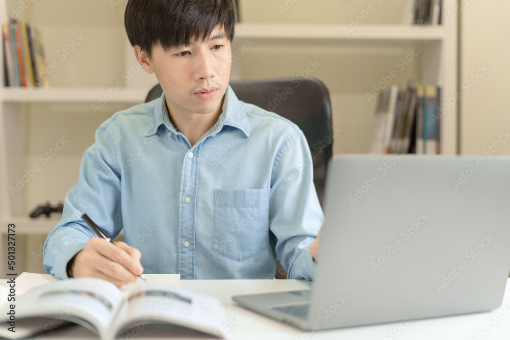 Young Asian college male student writing notes, using laptop for learning online education, business learning, watching online webinar. Focused man doing homework. Online education learning concept. Young Asian college male student writing notes, using laptop for learning online education, business learning, watching online webinar. Focused man doing homework. Online education learning concept.