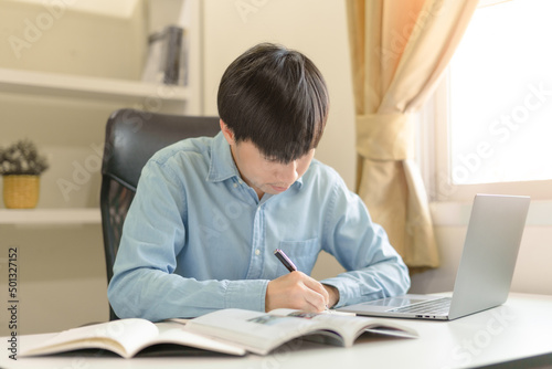 Young Asian college male student writing notes, using laptop for learning online education, business learning, watching online webinar. Focused man doing homework. Online education learning concept.