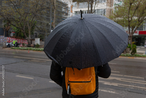 Back view of man withan umbrella in rainy day. He has orange backpack. Going school in bad weather.