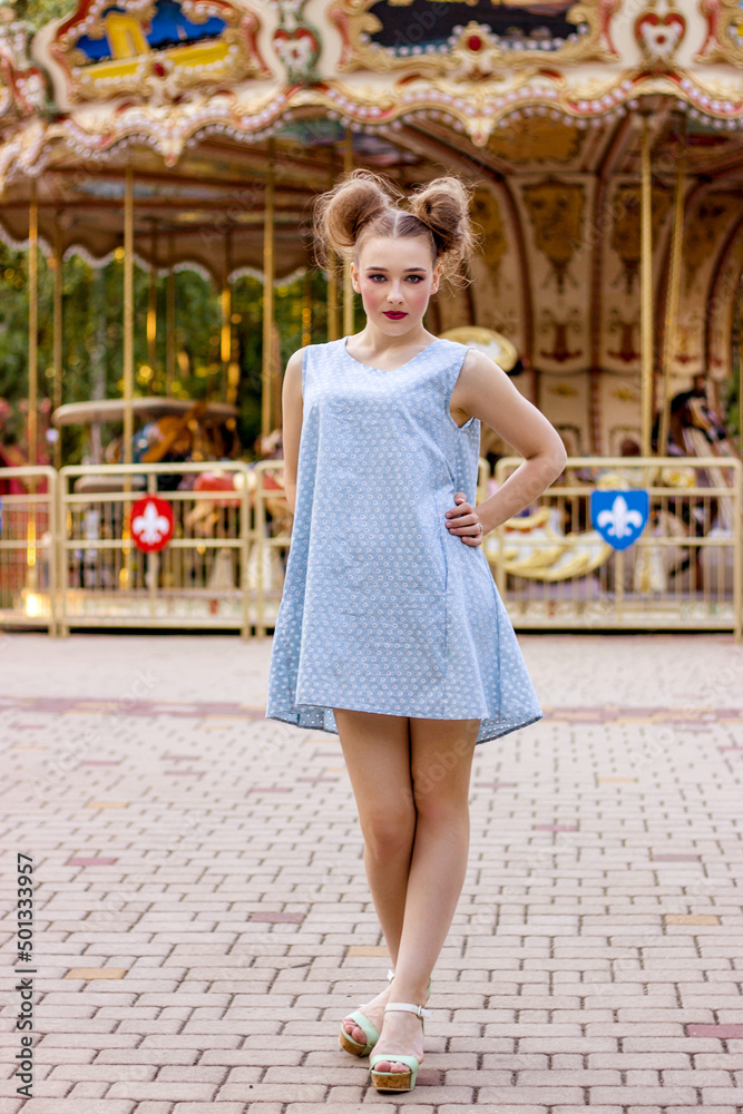A beautiful girl in a blue dress stands against the background of a carousel