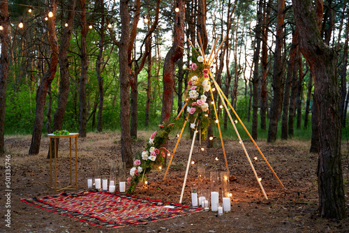 Bohemian tipi arch made of wooden rods decorated with pink roses  candles on carpet  pampass grass  wrapped in fairy lights on outdoor wedding ceremony venue in pine forest. Floristic compositions.