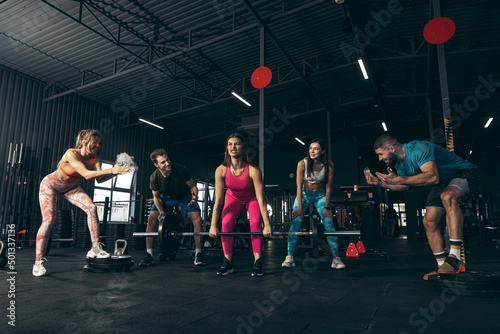 Group of young friends doing sports, training at gym indoors. Woman lifting barbell. Bulk up muscles