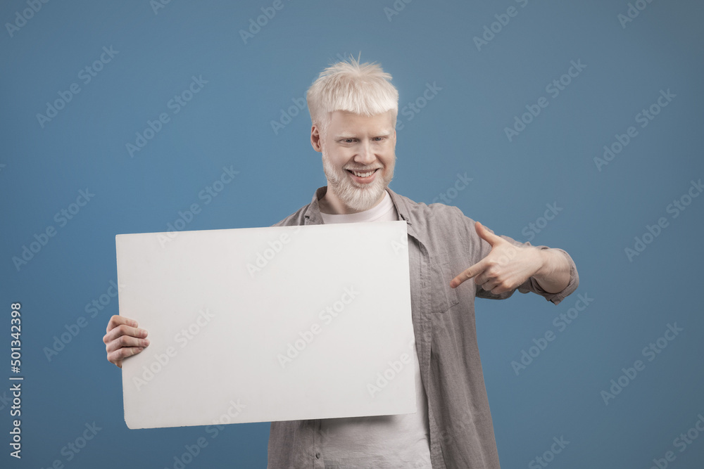 Place for your ad. Excited albino guy holding empty blank board and pointing at it, posing on blue background Place for your ad. Excited albino guy holding empty blank board and pointing at it, posing on blue background