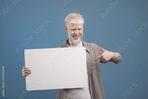 Place for your ad. Excited albino guy holding empty blank board and pointing at it, posing on blue background