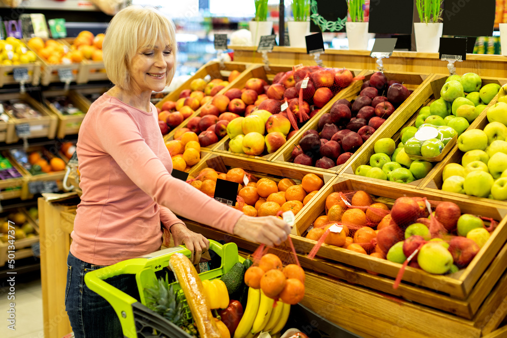 Positive elderly lady shopping at huge supermarket, buying fruits