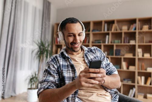 Handsome young middle eastern guy in wireless headphones listening to music online on smartphone at home