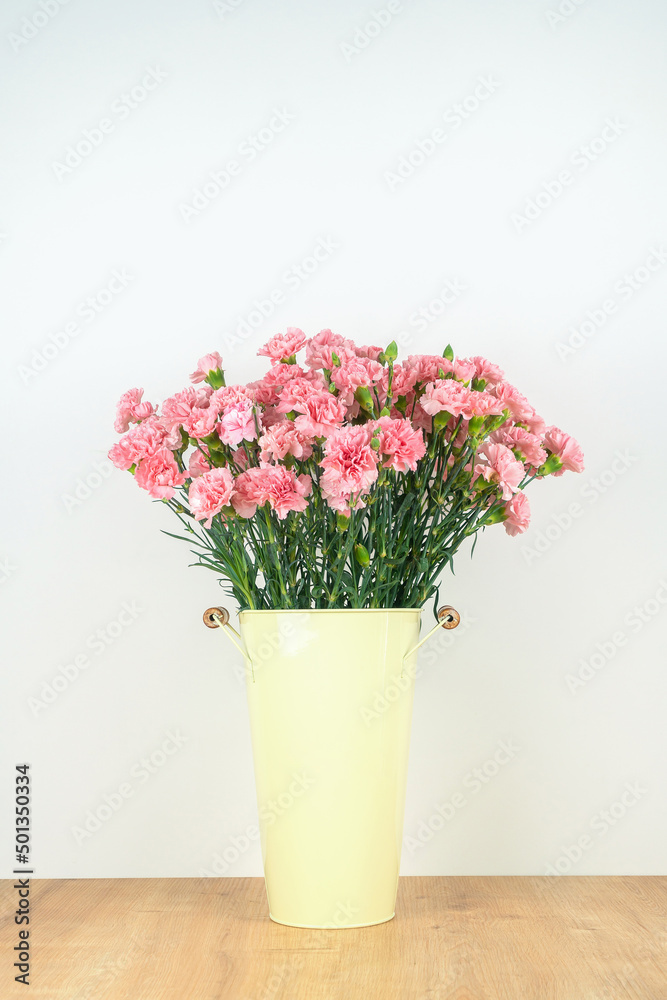 Close-up of a lot of pink carnations in a tin vase with handles on a wooden table on a neutral background. Vertical photo
