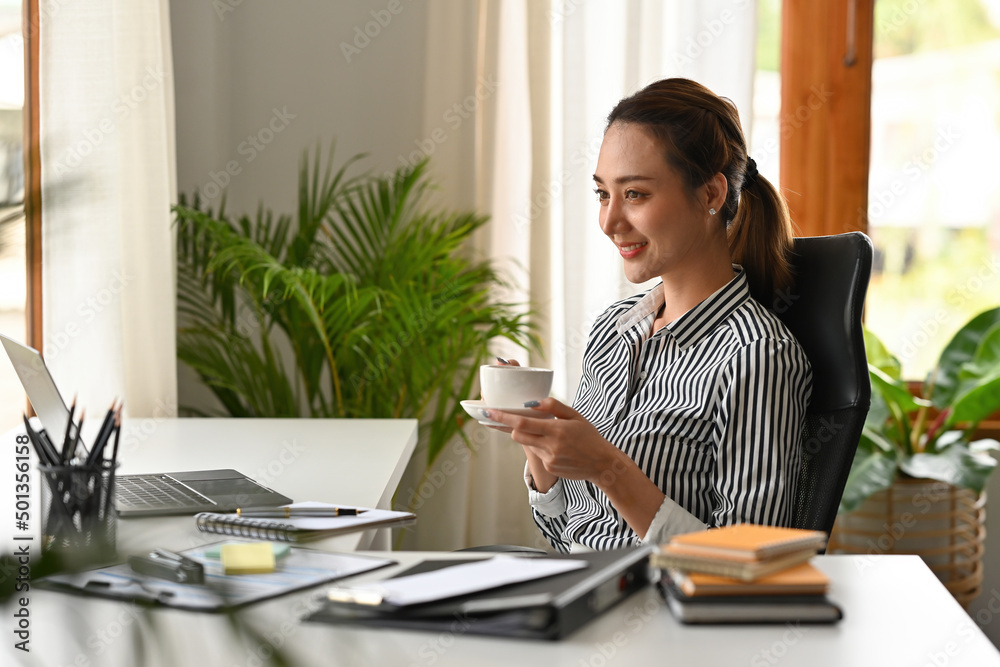 Side view of Indian lady enjoying morning coffee after writing a report in the modern office with the interior, business, and financial concepts Side view of Indian lady enjoying morning coffee after writing a report in the modern office with the interior, business, and financial concepts
