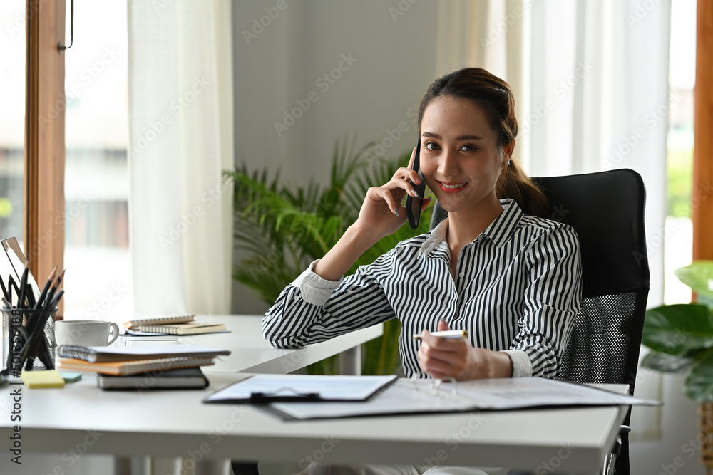 The portrait front view of a young Indian female smiling at the camera in a modern office while holding a pen in hand. Business and financial concepts. The portrait front view of a young Indian female smiling at the camera in a modern office while holding a pen in hand. Business and financial concepts.