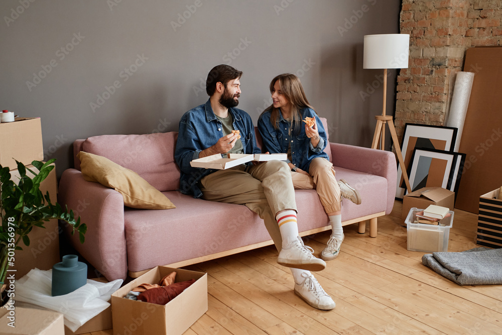 Modern young man and woman sitting together on sofa eating pizza in loft living room after moving to new apartment
