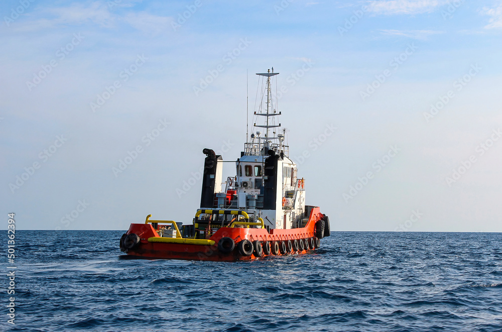 Red hulled tug boat with car tyres around it,  moving ahead in open waters. Rippled ocean surface and clear skies.