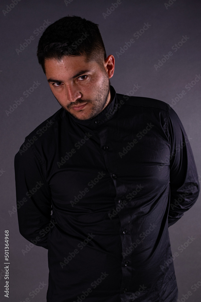 A man in a black shirt, posing on a gray background with his hands behind his back looking at the camera. Emotions, joy and fun of men.
