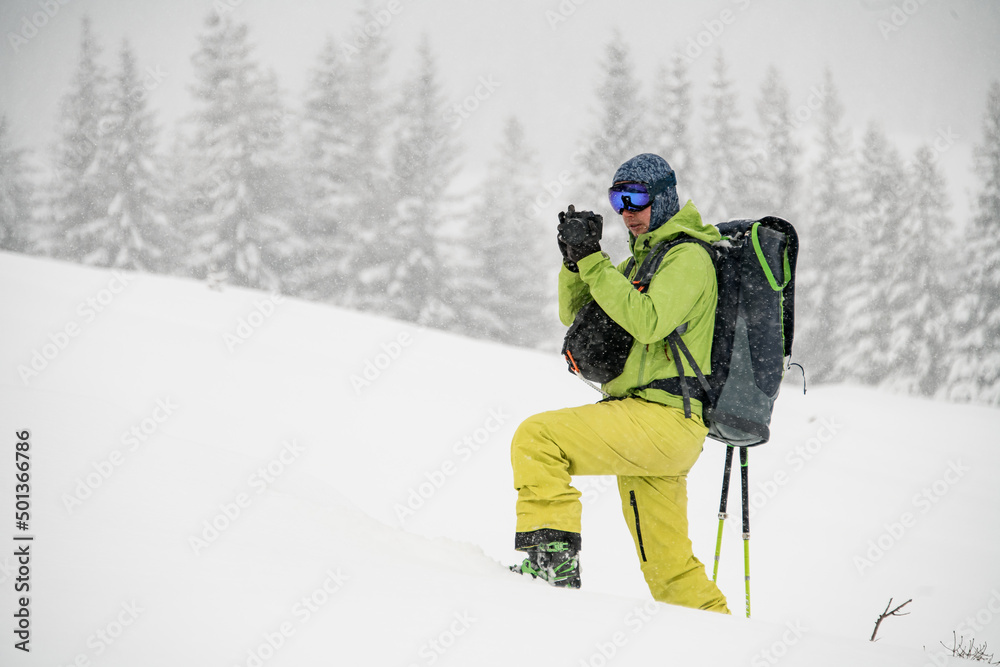 Side view of male skier in green and yellow ski suit with backpack standing alone and taking photo of snow-covered area