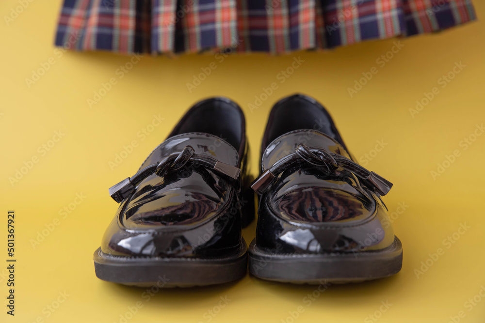 Black patent leather loafers and a plaid Scottish school skirt in the background on a yellow background