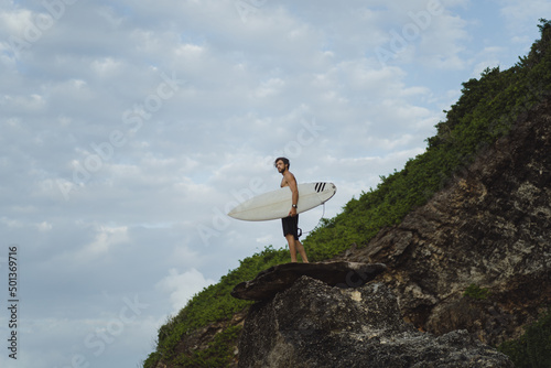 Young handsome man with a surfboard on a rock near the ocean.