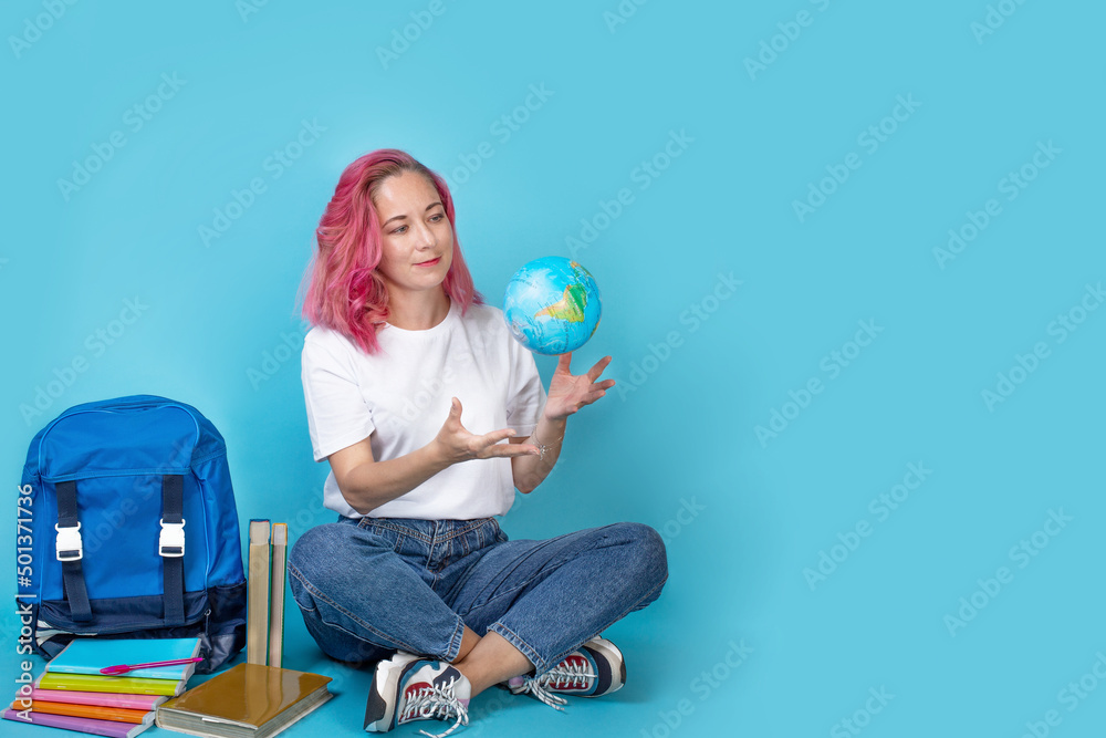 Young woman holding geographic globe of the world in her hands on blue background. Young female student sitting on floor with backpack and books. Long wide banner. education concept, copyspace Young woman holding geographic globe of the world in her hands on blue background. Young female student sitting on floor with backpack and books. Long wide banner. education concept, copyspace