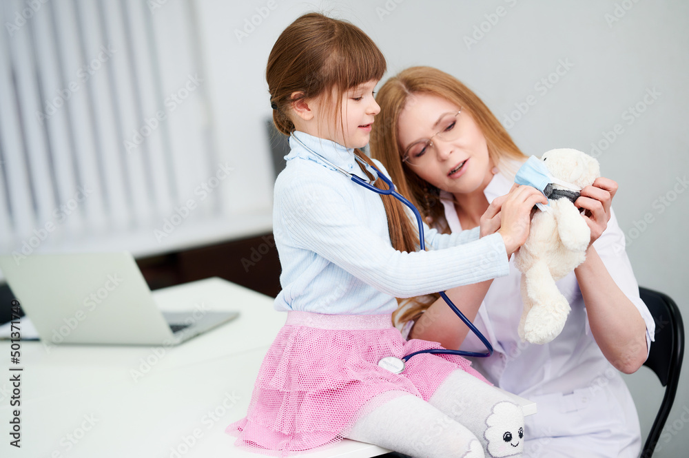 Obraz premium Caucasian female doctor with patient little girl examining by stethoscope bear toy in office