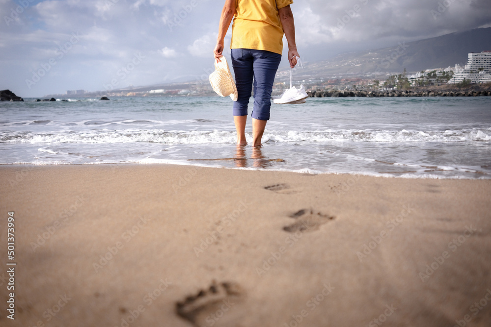 Rear view of senior woman barefoot on sea shore holding shoes and hat in hands while leaves footprints on the sand. Elderly woman enjoying vacation and freedom