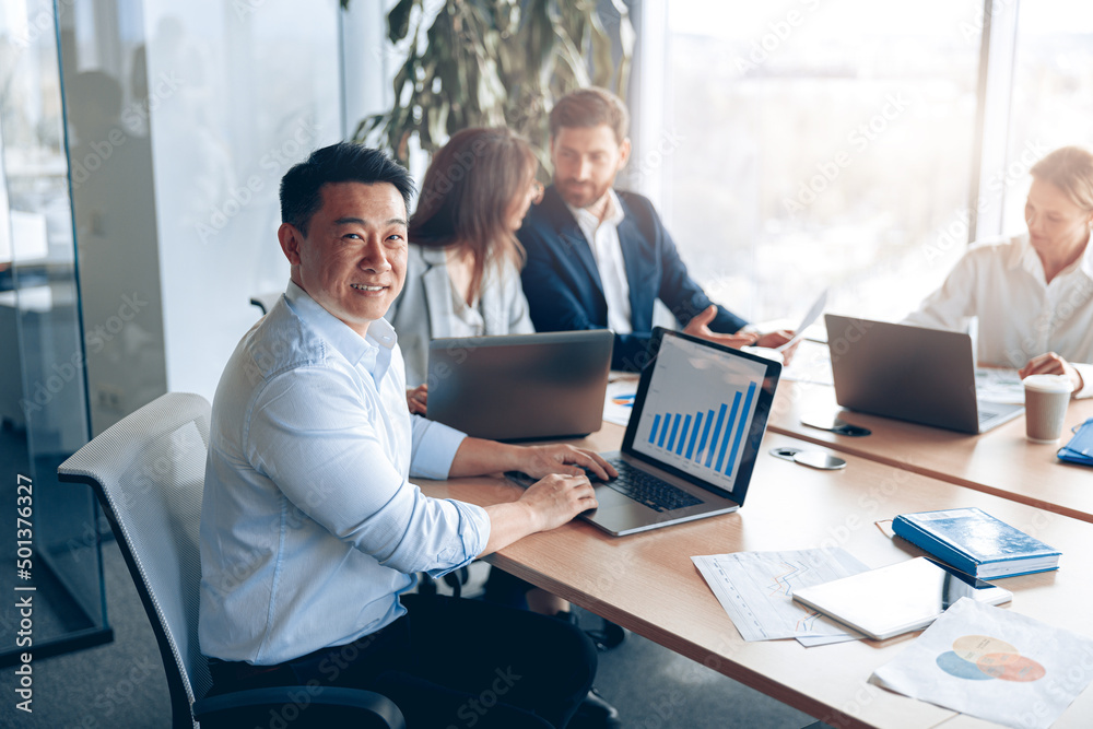 Portrait of smiling asian businessman working on a laptop during a meeting with a team. Business concept Portrait of smiling asian businessman working on a laptop during a meeting with a team. Business concept