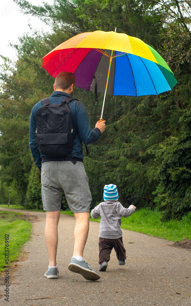 Not recognizable family in park. Father protecting a playing child from rain with a rainbow colored umrella, during a walk on rainy day.