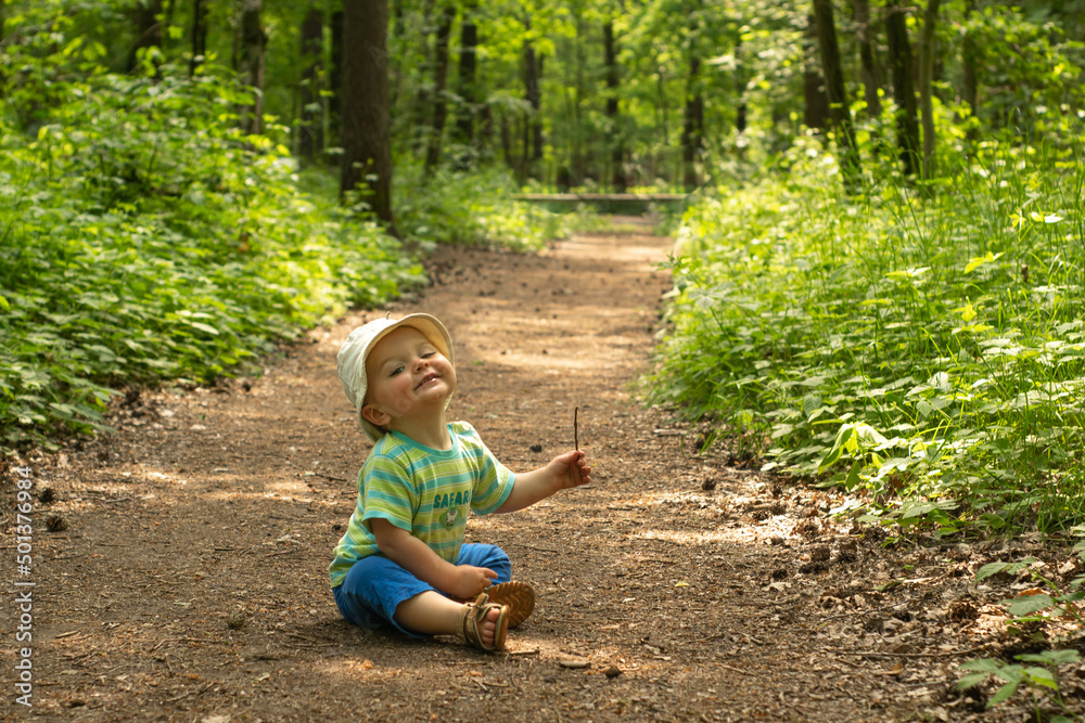 Tiny cute baby boy sitting and playing on the forest path. Child in summer clothes among nature during his countryside trip to the woods.