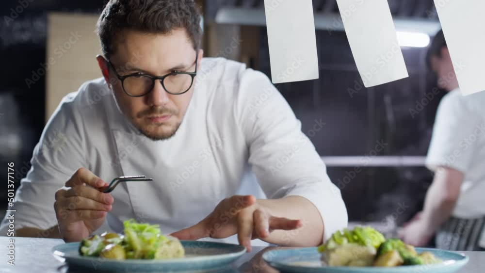 Professional male chef in glasses and uniform serving cooked dishes with tweezers on plates while working on food order in restaurant kitchen Professional male chef in glasses and uniform serving cooked dishes with tweezers on plates while working on food order in restaurant kitchen