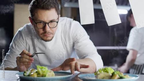 Professional male chef in glasses and uniform serving cooked dishes with tweezers on plates while working on food order in restaurant kitchen photo