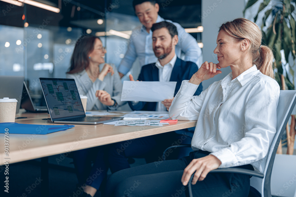 Smiling confident female boss sitting on meeting and looking at side with her colleagues at background in office. Business concept