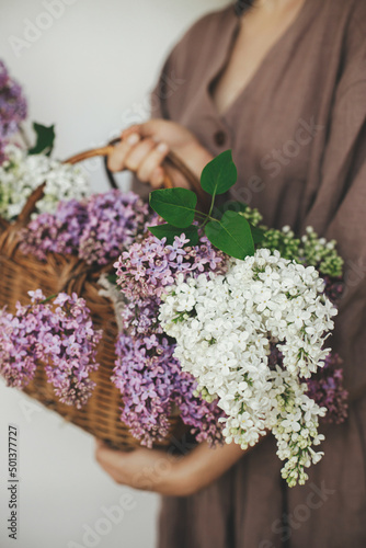 Stylish woman holding wicker basket with beautiful lilac flowers in rustic room. Female in linen dress arranging lilac flowers, cropped view. Authentic moody moment. Rustic wedding