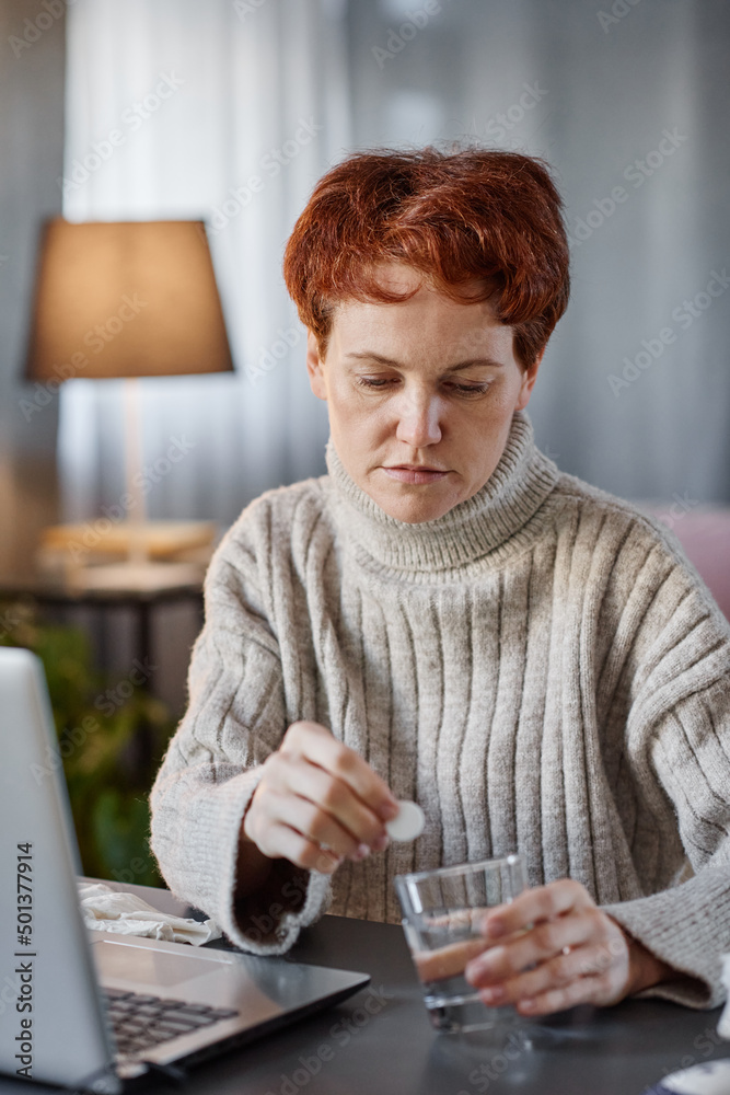 Vertical portrait of mature woman having influenza wearing sweater sitting at desk at home putting effervescent tablet into glass of water Vertical portrait of mature woman having influenza wearing sweater sitting at desk at home putting effervescent tablet into glass of water