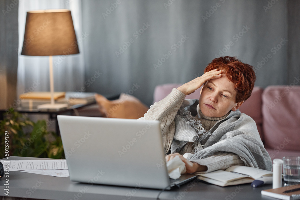 Portrait of mature Caucasian woman having flu with severe headache wearing warm outfit sitting at desk in living room at home trying to work Portrait of mature Caucasian woman having flu with severe headache wearing warm outfit sitting at desk in living room at home trying to work